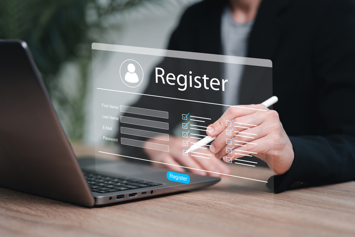 Person using a laptop while interacting with a digital registration form overlay labeled “Register,” entering details with a stylus, representing online account sign‑up or system registration.