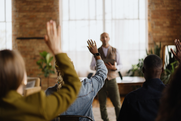 Audience members raising their hands while a speaker stands at the front of a room, presenting to a small group in a brick-walled workspace with large windows.