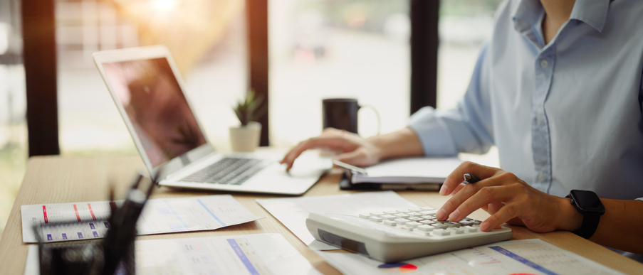 Person using a calculator at a desk with financial documents spread out and an open laptop in the background, representing budgeting or tax calculations.